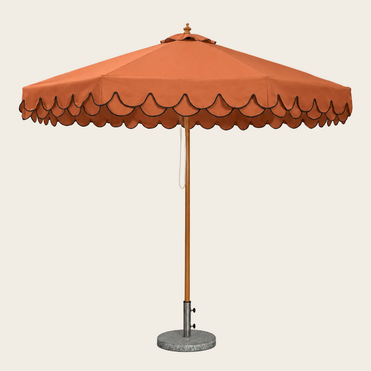 An orange patio umbrella with a scalloped edge, a wooden pole, and a round concrete base on a white background.