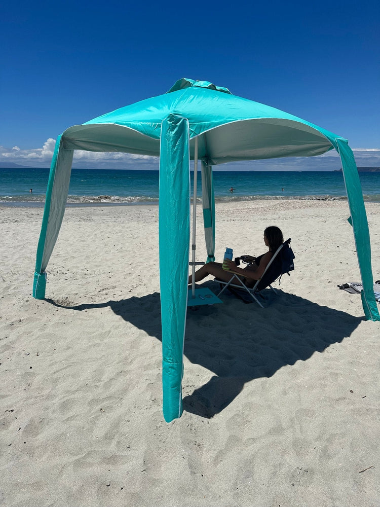 Turquoise Ultimate Beach Cabana providing UPF50+ shade on a sunny beach with a person relaxing underneath