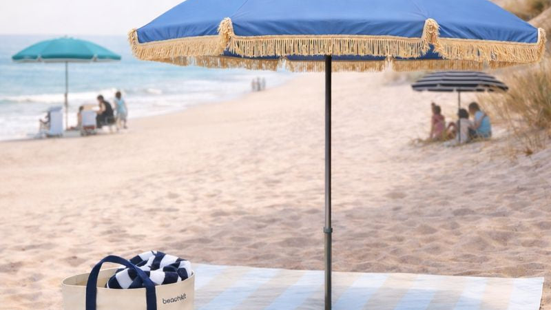 Beach scene with a blue umbrella, striped towel, and cooler bag on sand.