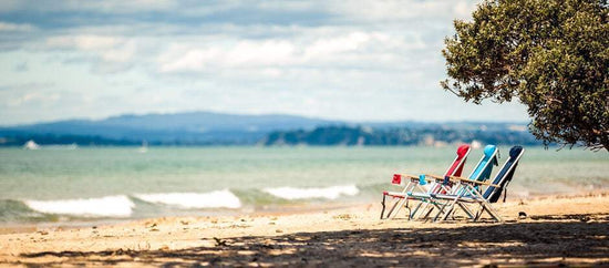Three Beachkit Wearever Backpack Chairs in red, blue, and black on a sandy beach with ocean and distant mountains in the background
