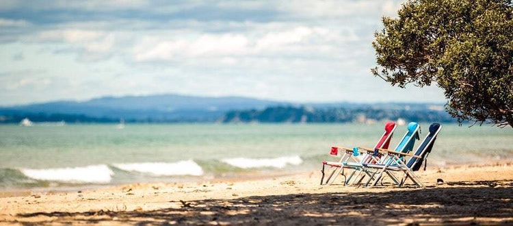 Three Beachkit Wearever Backpack Chairs in red, blue, and black on a sandy beach with ocean and distant mountains in the background